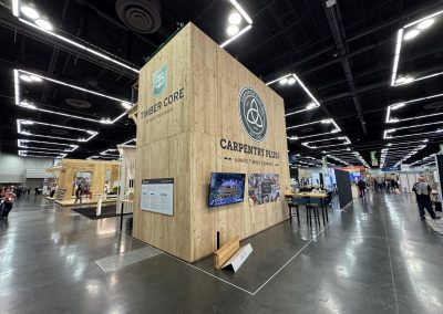 A large Carpentry Plus trade show booth with timber walls, display screens, and informational panels inside a spacious convention center.